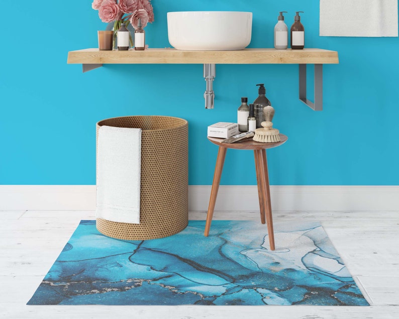 Puede incluir: Un ba&ntilde;o con una pared azul, un estante de madera con un lavabo blanco, una cesta de lavander&iacute;a de mimbre con una toalla blanca, una peque&ntilde;a mesa de madera con art&iacute;culos de tocador y una alfombra de ba&ntilde;o con estampado abstracto azul y blanco.