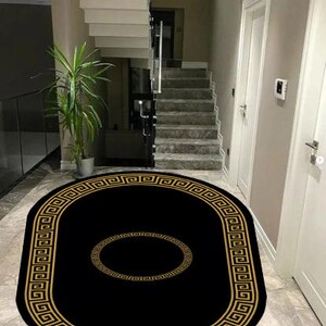 Puede incluir: Una alfombra negra de forma ovalada que presenta un borde dorado con un patr&oacute;n de greca a lo largo del contorno y un motivo circular dorado a juego centrado en el medio, creando un contraste llamativo sobre el fondo oscuro de la pieza.