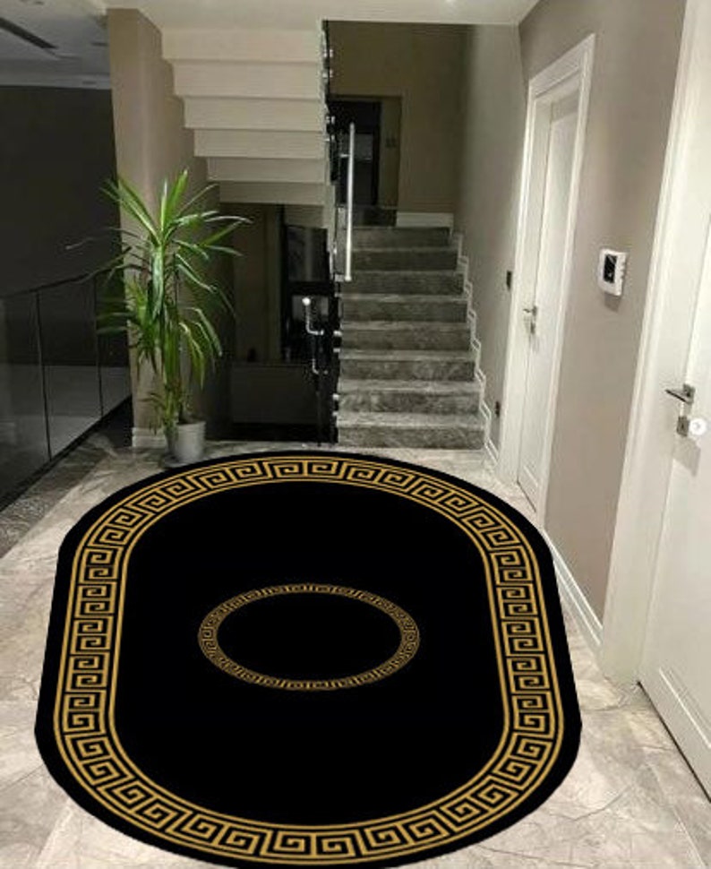 Puede incluir: Una alfombra negra de forma ovalada que presenta un borde dorado con un patr&oacute;n de greca a lo largo del contorno y un motivo circular dorado a juego centrado en el medio, creando un contraste llamativo sobre el fondo oscuro de la pieza.