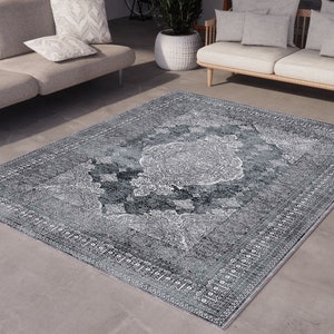 May include: A grey and white patterned area rug with a vintage design. The rug is laid out on a grey concrete floor in front of a light grey sofa.