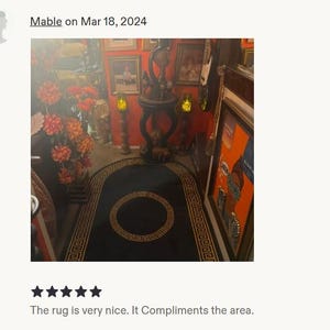 Puede incluir: Una alfombra negra con un borde dorado de patr&oacute;n de greca y un motivo circular central. La alfombra es rectangular con extremos redondeados y est&aacute; colocada en el suelo de una habitaci&oacute;n con paredes naranjas, muebles y decoraci&oacute;n floral.