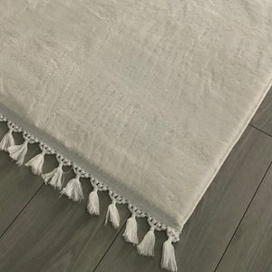 May include: White fluffy rug with a white fringe border. The rug is lying on a wooden floor.