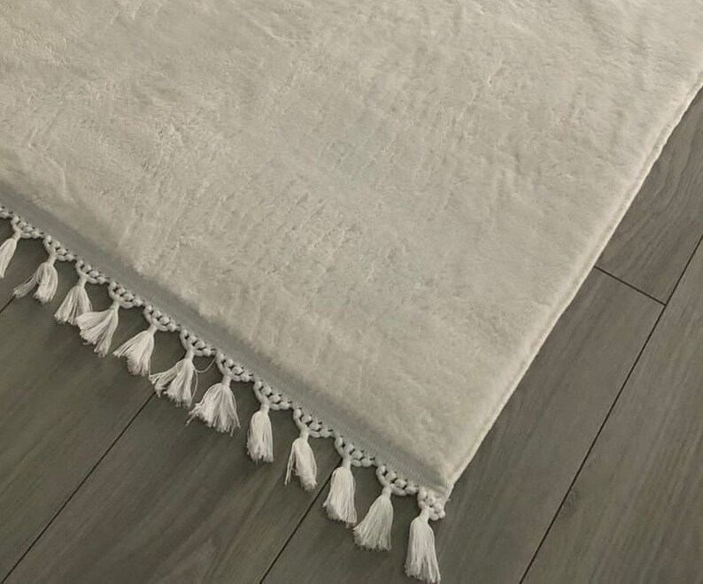 May include: White fluffy rug with a white fringe border. The rug is lying on a wooden floor.