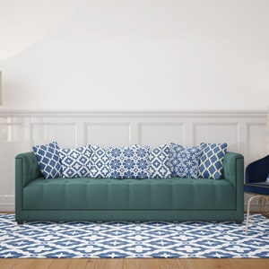 Puede incluir: Un sof&aacute; verde azulado con cinco cojines decorativos con patrones azules y blancos. El sof&aacute; est&aacute; en una sala de estar con una alfombra blanca con un patr&oacute;n geom&eacute;trico azul y blanco, una mesa auxiliar blanca y una l&aacute;mpara con una pantalla beige.