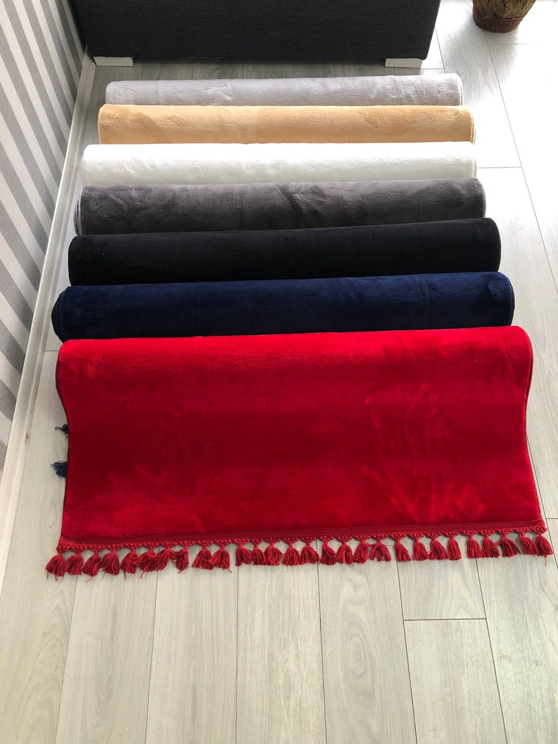 May include: A stack of rolled-up rugs in various colours, including red, navy blue, black, grey, white, beige, and light grey. The rugs have a fringe trim.
