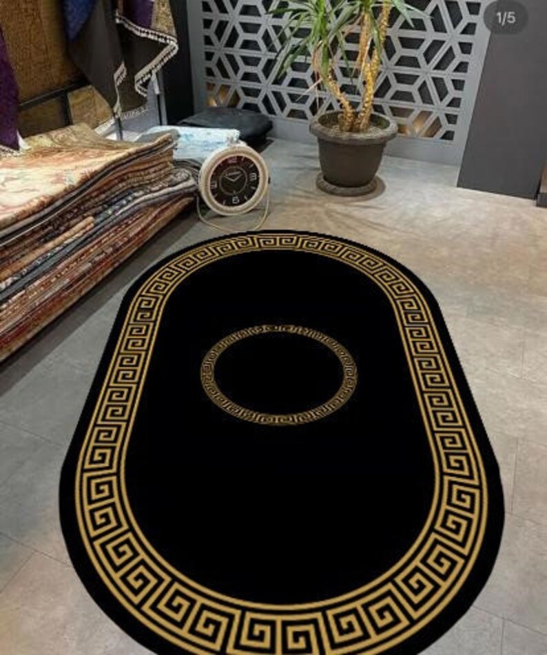 Puede incluir: Una alfombra negra de forma ovalada que presenta un patr&oacute;n de borde de greca dorada a lo largo del borde exterior y un dise&ntilde;o circular de greca dorada a juego centrado en el medio de la alfombra. La alfombra se muestra sobre un suelo gris claro.