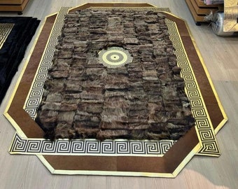 Puede incluir: Una alfombra marrón y dorada con un patrón geométrico. La alfombra tiene un medallón central con un diseño de llave griega. La alfombra está hecha de piel.