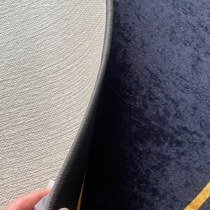 Puede incluir: Una alfombra circular con una superficie de terciopelo azul marino oscuro y l&iacute;neas geom&eacute;tricas doradas. El reverso es de material texturizado beige claro con un borde cosido en gris oscuro y una peque&ntilde;a etiqueta de cuidado blanca.