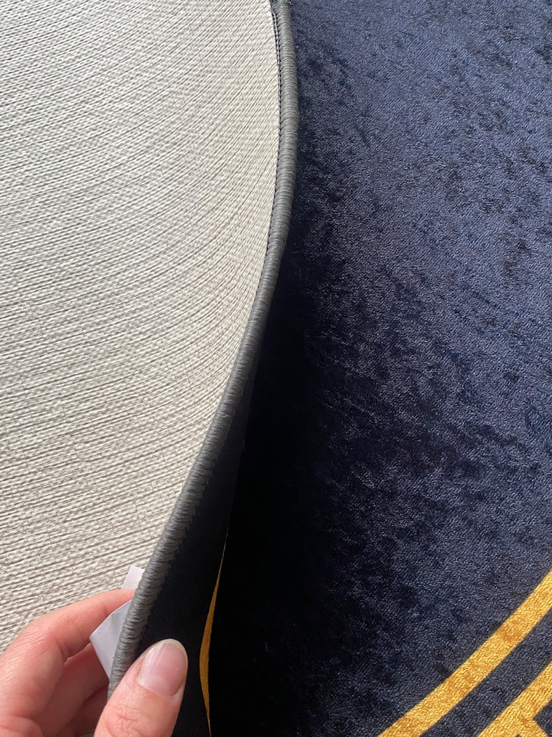 Puede incluir: Una alfombra circular con una superficie de terciopelo azul marino oscuro y l&iacute;neas geom&eacute;tricas doradas. El reverso es de material texturizado beige claro con un borde cosido en gris oscuro y una peque&ntilde;a etiqueta de cuidado blanca.