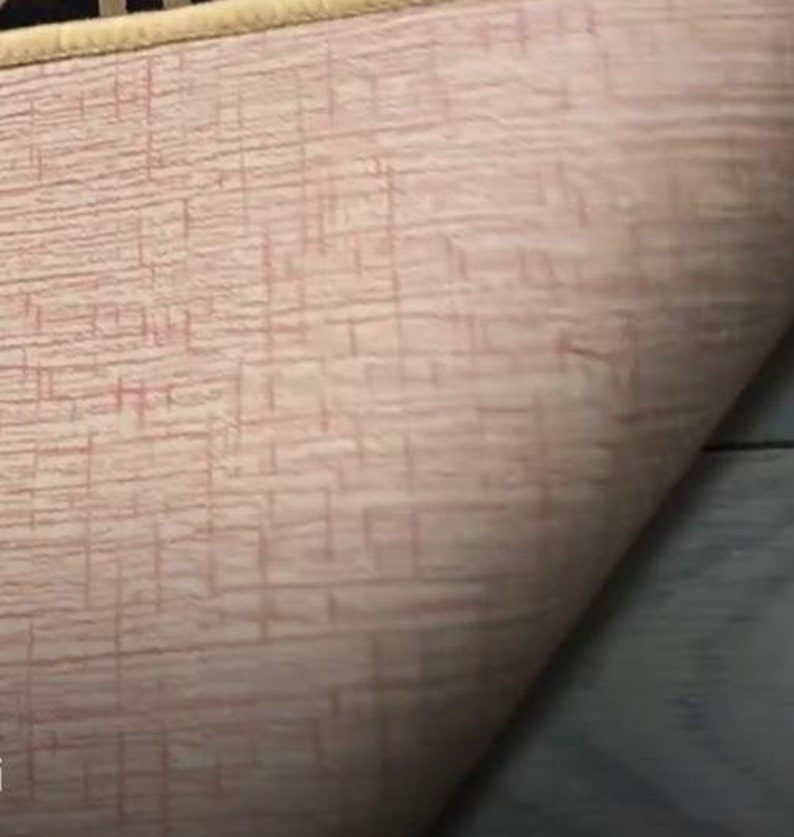 May include: A light pink fabric with a textured, grid-like pattern of thin, darker pink lines. The material has a soft, slightly fuzzy appearance and features a distinct, rounded edge finished with a light beige or cream-colored trim.