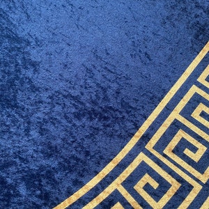 Puede incluir: Una tela azul profundo con textura de terciopelo arrugado que presenta un prominente patr&oacute;n de llave griega de color dorado que corre diagonalmente por la esquina inferior derecha. El material muestra un brillo rico con tonos variados.