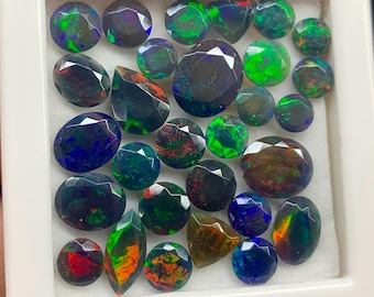 May include: A collection of 25 loose, faceted, iridescent black opals in various shapes and sizes. The opals are displayed in a white box with a black border.
