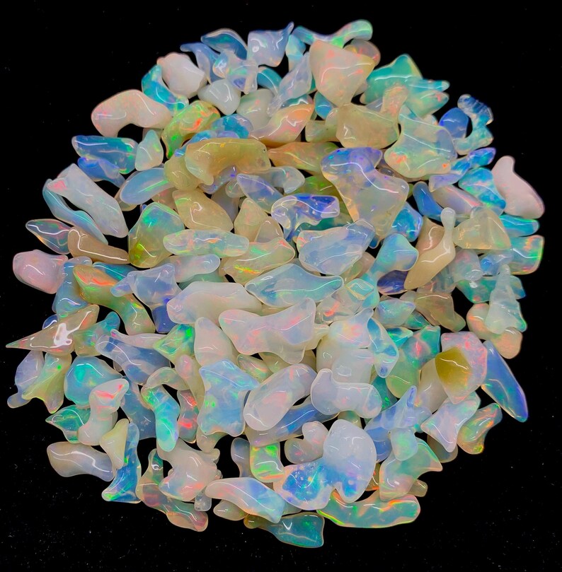 以下が含まれることがあります： 様々な形と大きさの虹色のオパールチップが大量に積み重ねられています。チップは主に白で、青、緑、黄色の光沢があります。