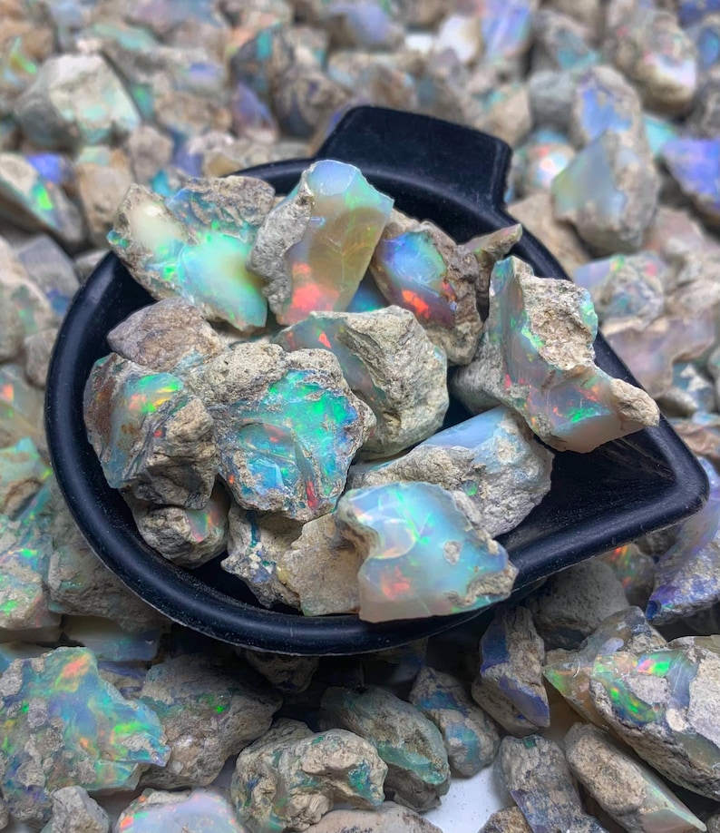 Op de afbeelding: Een zwarte schep gevuld met ruwe opaal edelstenen, met levendige kleurflitsen. De opalen tonen een mix van blauw, groen en rood, tegen een achtergrond van andere ruwe opaalstenen. De opalen hebben een ruwe, natuurlijke textuur.