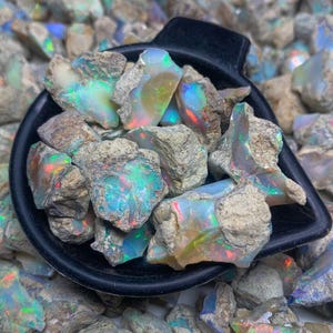 Op de afbeelding: Een zwarte schep vol ruwe opaal edelstenen, met levendige iriserende kleuren van groen, blauw en rood. De opalen staan tegen een achtergrond van vergelijkbare ruwe stenen.