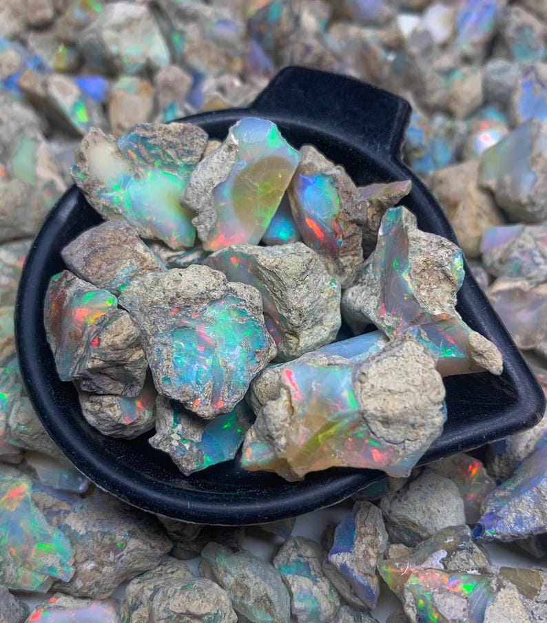 Op de afbeelding: Een zwarte schep vol ruwe opaal edelstenen, met levendige iriserende kleuren van groen, blauw en rood. De opalen staan tegen een achtergrond van vergelijkbare ruwe stenen.