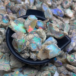 Op de afbeelding: Een zwarte lepel vol ruwe opaal edelstenen, met levendige kleurflitsen. De opalen tonen een mix van blauw, groen en rood, tegen een neutrale, rotsachtige achtergrond. De edelstenen zijn ruw en ongeslepen.