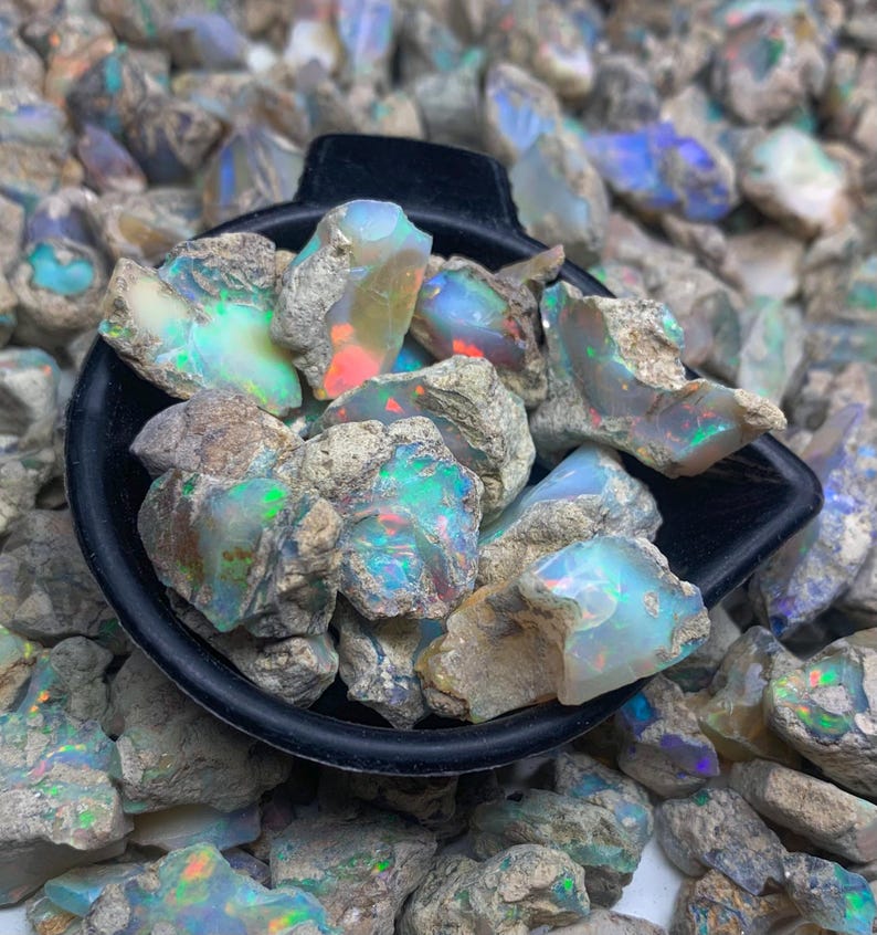 Op de afbeelding: Een zwarte lepel vol ruwe opaal edelstenen, met levendige kleurflitsen. De opalen tonen een mix van blauw, groen en rood, tegen een neutrale, rotsachtige achtergrond. De edelstenen zijn ruw en ongeslepen.