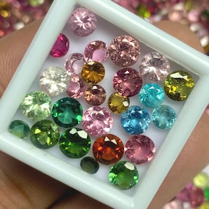 May include: A selection of multi-coloured, faceted gemstones, including pink, green, blue, and orange, presented in a white square tray. The gemstones are round and of different sizes, with a sparkling appearance.