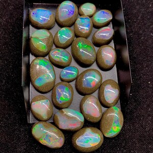 May include: A collection of 20 oval-shaped opal gemstones in various shades of green, blue, and white. The opals are set in a dark grey matrix and are displayed in a black box.
