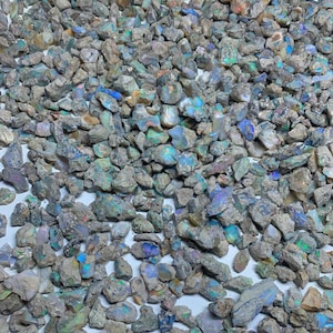 Op de afbeelding: Een verzameling ruwe opaalstenen, met een verscheidenheid aan kleuren, waaronder blauw, groen en iriserende tinten. De stenen hebben een natuurlijke, onbewerkte uitstraling, met verschillende maten en vormen. Het kleurenpalet is een mix van neutrale grijstinten en aardetinten.