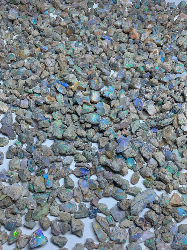 Op de afbeelding: Een verzameling ruwe opaalstenen, met een verscheidenheid aan kleuren, waaronder blauw, groen en iriserende tinten. De stenen hebben een natuurlijke, onbewerkte uitstraling, met verschillende maten en vormen. Het kleurenpalet is een mix van neutrale grijstinten en aardetinten.