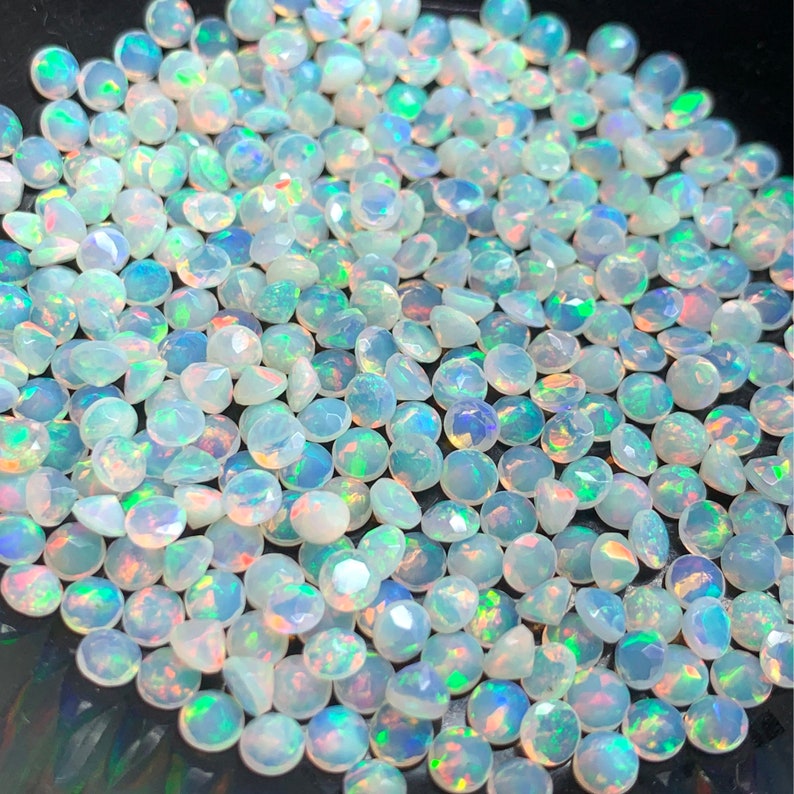 May include: A large group of iridescent white opal gemstones, each with a round, faceted shape. The opals are scattered on a black surface.