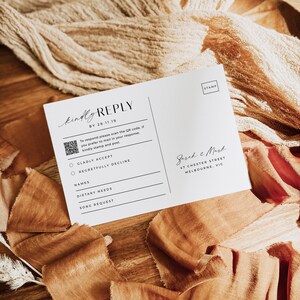 May include: A white RSVP card with a black border and the text "kindly REPLY BY 28.11.19". The card has a QR code and two options for guests to respond: "GLADLY ACCEPT" or "REGRETFULLY DECLINE". The card also includes spaces for guests to write their names, dietary needs, and song requests. The card is addressed to "Sarah & Mark" with their address: "47 Chester Street, Melbourne, VIC".