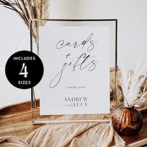 May include: A white sign with black calligraphy that reads "cards + gifts" and "Thank You" with the names "Andrew and Lucy" below. The sign is in a gold frame and sitting on a wooden table with a brown vase and dried flowers.
