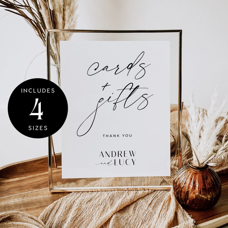 May include: A white sign with black calligraphy that reads "cards + gifts" and "Thank You" with the names "Andrew and Lucy" below. The sign is in a gold frame and sitting on a wooden table with a brown vase and dried flowers.