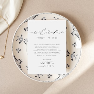 May include: A white welcome sign with black text for a wedding reception. The text reads "Welcome Family + Friends" and includes a message thanking guests for sharing in the joy of the wedding day. The sign is signed "With Love Andrew and Lucy".