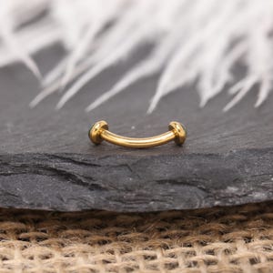 May include: A curved, gold-colored eyebrow ring with a small, round, dark green gem on each end. The jewelry is displayed on a dark gray, textured surface. The background features blurred white feathers.