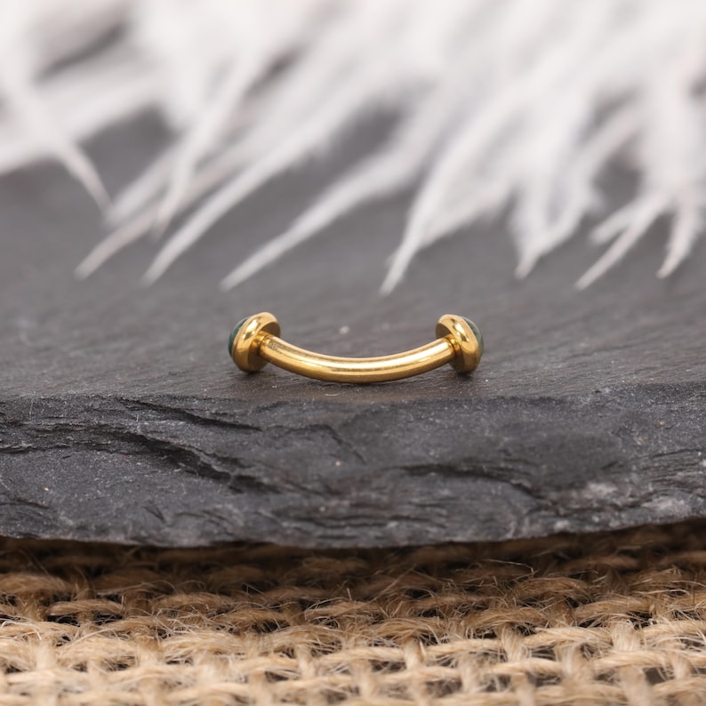 May include: A curved, gold-colored eyebrow ring with a small, round, dark green gem on each end. The jewelry is displayed on a dark gray, textured surface. The background features blurred white feathers.