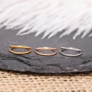 May include: Three minimalist rings in gold, rose gold, and silver. Each ring features a double-band design, with a smooth, polished finish. The rings are displayed on a dark gray surface, with a blurred white background.