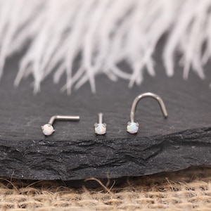 Könnte beinhalten: Drei Nasenpiercings mit einem kleinen, runden, weißen Opalstein. Das Set beinhaltet einen geraden Stecker, einen gebogenen Stecker und einen L-förmigen Stecker. Der Schmuck ist vor einem dunkelgrauen Hintergrund mit weißen Federn platziert.