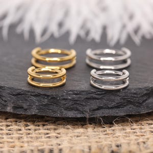 Könnte beinhalten: Goldene und silberne Ohrklemmen mit Doppelband. Die Ohrklemmen werden auf einer dunkelgrauen Oberfläche präsentiert. Die goldenen Ohrklemmen haben einen warmen, hellen Ton, während die silbernen einen kühlen, metallischen Glanz aufweisen. Das Design ist schlicht und modern.