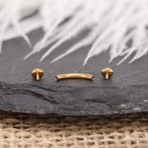 May include: A gold-colored curved barbell body piercing jewelry with two round, screw-on ends. The jewelry is displayed on a dark gray surface with a blurred white background.
