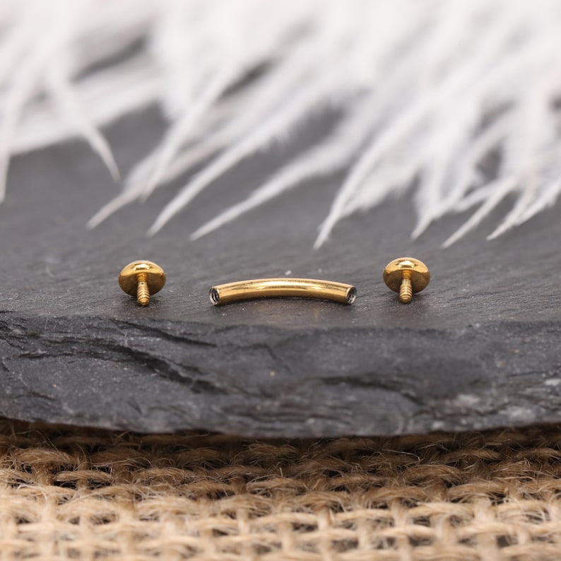 May include: A gold-colored curved barbell body piercing jewelry with two round, screw-on ends. The jewelry is displayed on a dark gray surface with a blurred white background.