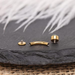 May include: Gold-coloured body jewellery components, including a curved barbell, a flat disc, and a threaded end with a small, round, green gem. The pieces are arranged on a dark, textured surface.