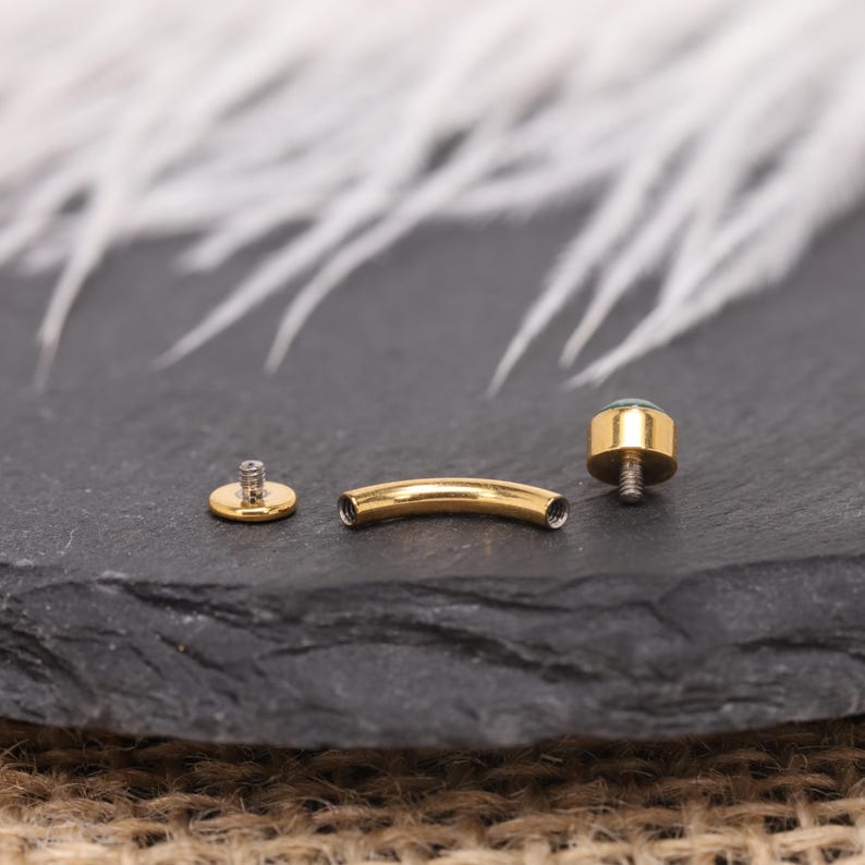 May include: Gold-coloured body jewellery components, including a curved barbell, a flat disc, and a threaded end with a small, round, green gem. The pieces are arranged on a dark, textured surface.