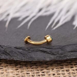 May include: A curved, gold-coloured barbell-style piercing stud with a small, round, teal-coloured stone on one end. The jewellery is displayed on a dark grey, textured surface.