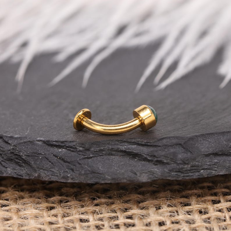 May include: A curved, gold-coloured barbell-style piercing stud with a small, round, teal-coloured stone on one end. The jewellery is displayed on a dark grey, textured surface.