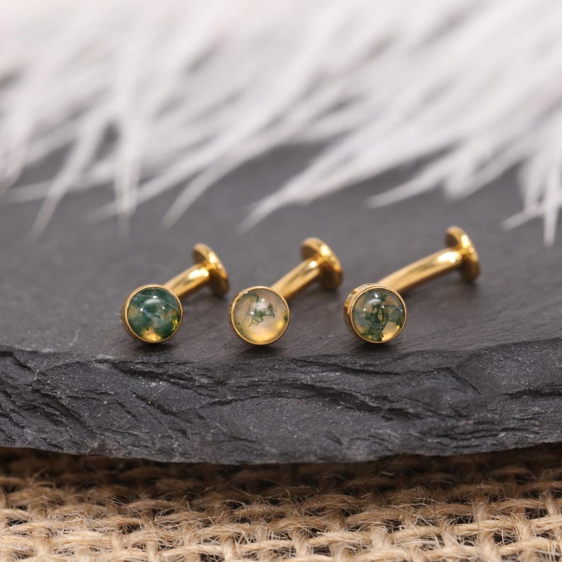 May include: Three gold-coloured labret studs with round moss agate stones. The stones have green and white inclusions. The studs are displayed on a dark grey surface, with a blurred white background.