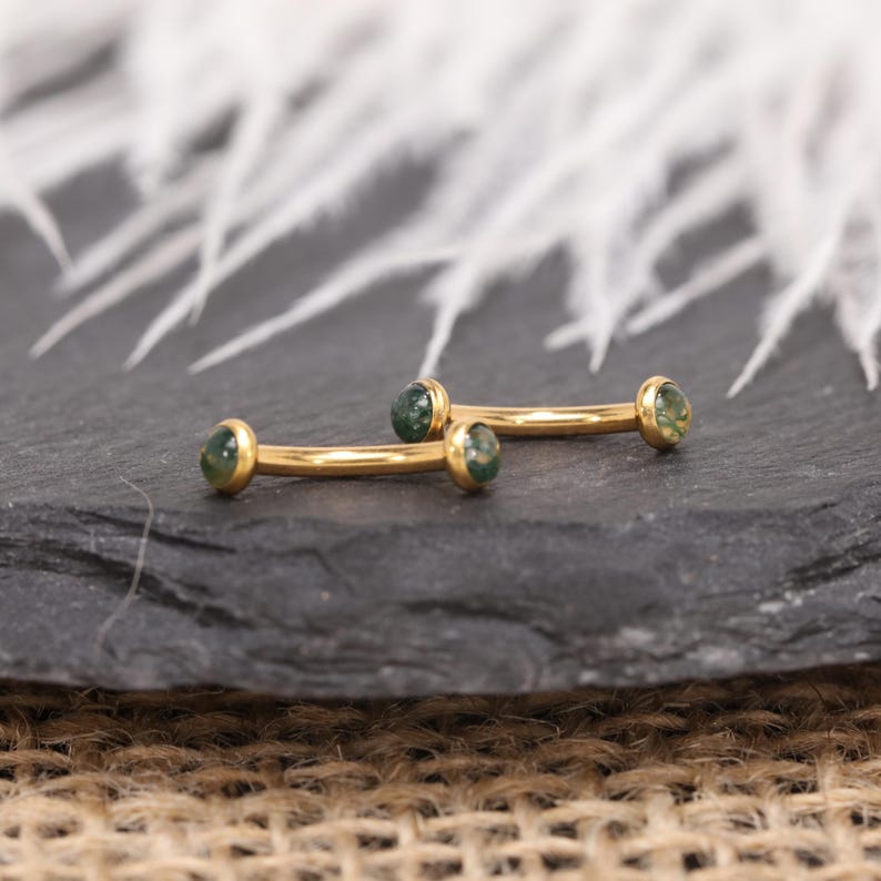 May include: Two gold-colored curved barbells with green gemstone ends. The jewelry is displayed on a dark gray surface, with a blurred white feather-like background. The barbells are designed for body piercings.