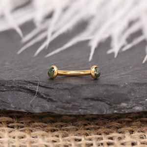 May include: A gold-colored barbell-style piercing with green gemstone ends. The curved bar is centered on a dark gray surface, with white feathers in the background. The jewelry is designed for body piercings.
