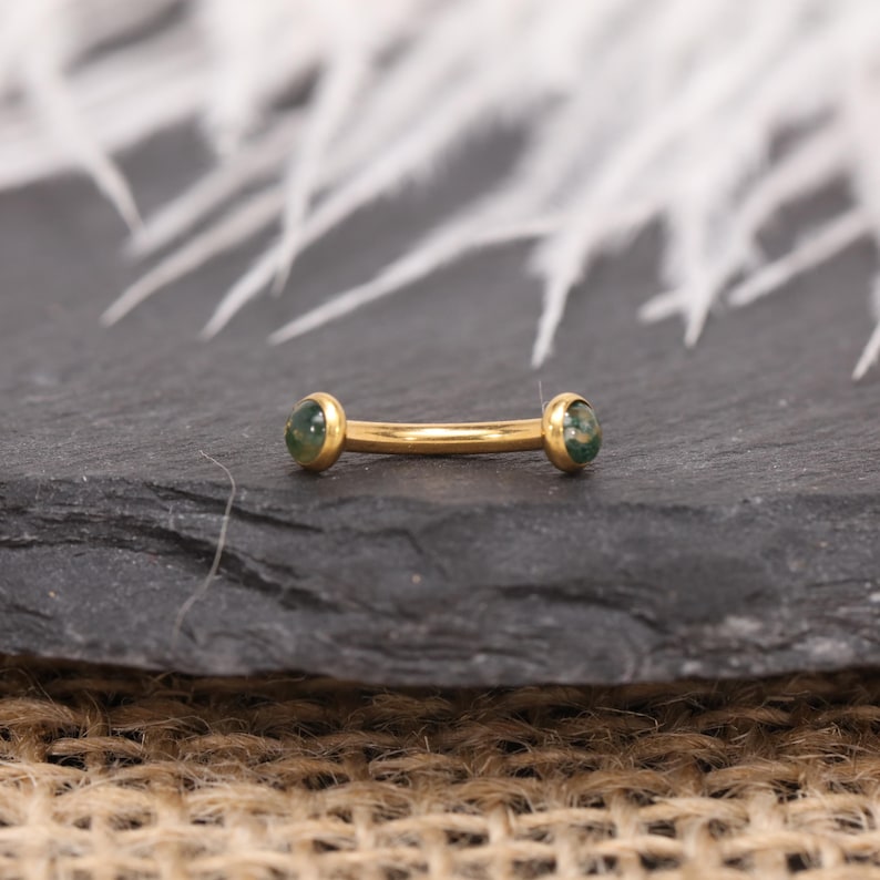 May include: A gold-colored barbell-style piercing with green gemstone ends. The curved bar is centered on a dark gray surface, with white feathers in the background. The jewelry is designed for body piercings.