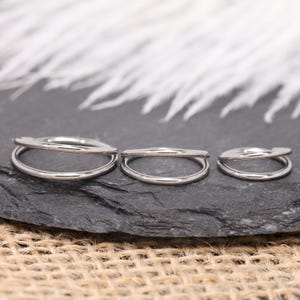 May include: Three silver-colored, double-hoop rings of varying sizes are displayed on a dark gray, textured surface. The rings are arranged in a row, with the largest on the left and the smallest on the right. The background features white, feathery elements.