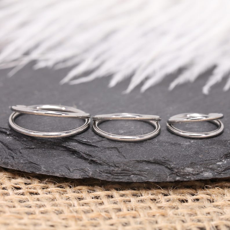 May include: Three silver-colored, double-hoop rings of varying sizes are displayed on a dark gray, textured surface. The rings are arranged in a row, with the largest on the left and the smallest on the right. The background features white, feathery elements.