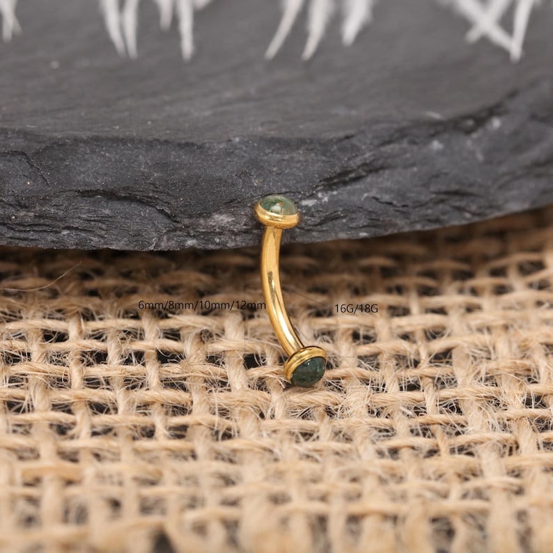 May include: A curved eyebrow piercing with a gold-colored bar and two green gemstones. The text on the image indicates sizes of 6mm, 8mm, 10mm, and 12mm, and gauge sizes 16G/18G. The jewelry is displayed on a textured surface.