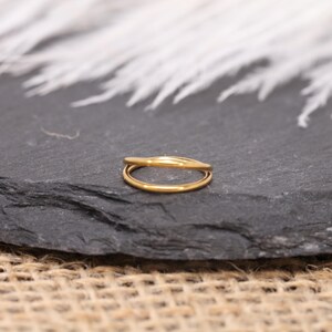 May include: A gold-colored ring with a double-band design. The ring is made of a shiny, reflective metal and sits on a dark gray surface. The ring's bands are smooth and rounded, creating a minimalist and elegant look.